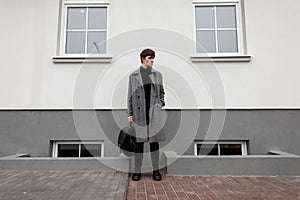 European urban young man in stylish autumn-winter outerwear with a fashionable leather black backpack in trendy boots stands on