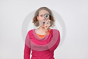 European woman in pink sweater looking through a magnifying glass with opened surprised mouth.