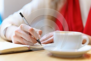 European woman with red scarf is writing by pen somthing in the notepad near white cup of coffee on table