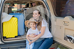 European woman sitting with her son in the trunk of an SUV. Beautiful happy mother loving hugs son. Yellow luggage for