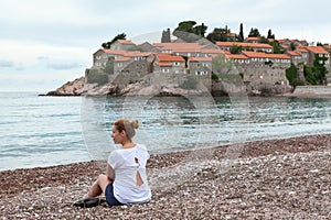 European woman sitting on the luxury beach against Sveti Stefan hotel and resort. Adriatic sea, Montenegro. Europa