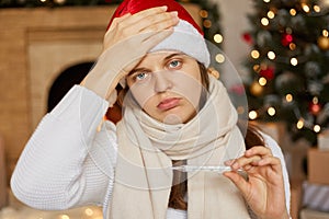 European woman with very high body temperature, young sick woman in red santa and scarf measures the temperature with thermometer