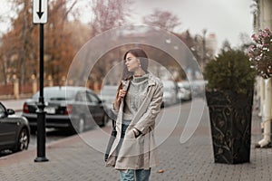 European young brown-haired woman in a sweater in a fashionable long light trench coat with a black leather bag posing outdoors on