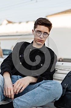 European young hipster man in trendy casual denim clothes in stylish glasses with trendy black backpack enjoys relaxation and