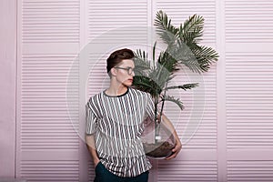 European young man with a fashionable hairstyle in stylish youth clothes in vintage glasses with a green plant is resting near a