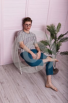 European young man with a fashionable hairstyle in stylish youth clothes in vintage glasses is resting on a chair next to a green