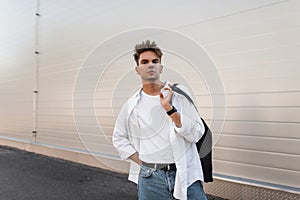 European young man with a stylish hairstyle in a trendy white shirt in vintage jeans is standing near a modern white building