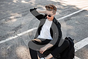 European young man with trendy hairstyle in elegant clothes in sunglasses with a backpack sits on a rubber wheel outdoors