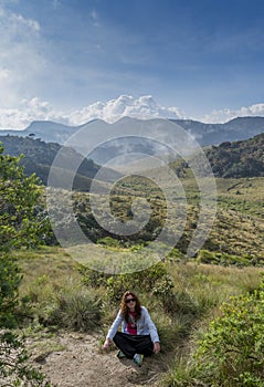 European young tourist girl sitting on the grass at the beautiful valley surrounded by mountains