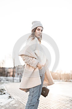 European young woman in a fashionable milk-colored fur faux coat in vintage jeans in a knitted trendy hat with a leather handbag