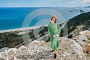 European young woman in green summer dress poses for photographer on rock above sea in early spring Antalya, Turkey.
