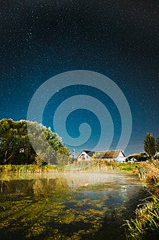 Evaporation Over River Lake Near Houses In Village. Milky Way Galaxy In Night Starry Sky Above Lake River Landscape At