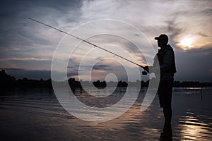 It is evening outside. Fisherman stands in shallow and fishing. He holds fly rod in hands. There is a reel under it. He