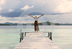 An evening shot of a tourist woman standing with oper arms, hat, at the end of a pier looking in the water and at the sunset