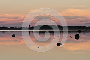 Evening sunlight, lighthouse and rocks on coast. Sky reflection on water. Beach in summer. Seaside natural environment.