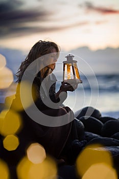 Evening after sunset at the beach, blonde woman sits with lantern beside the sea, light bokeh