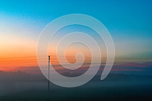 Evening thick fog over field and forest. Landscape of the evening sky with orange and blue sky and fog. Telegraph pole in the fog.