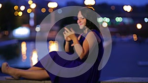 Evening time a young woman in a blue dress is sitting on a steamer in the street reading a message in a smartphone smiling and dre