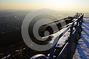 Večerný pohľad na panorámu mesta z vyhliadkového kopca s oceľovou koľajnicou a starou lavicou počas zasneženej sezóny. Lokalita Nitra mesto, Slovensko