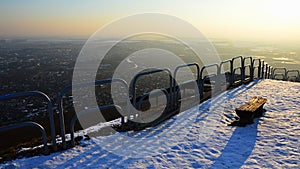 Večerný pohľad na panorámu mesta z vyhliadkového kopca s oceľovou koľajnicou a starou lavicou počas zasneženej sezóny. Lokalita Nitra mesto, Slovensko