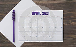 Event planner for April 2021. Calendar page on white paper, next to purple pencil. Brown wooden background. Table with the days
