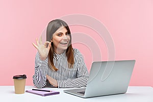 Everything is fine! Positive satisfied businesswoman in striped shirt showing ok gesture, approval hand sign, working on laptop at