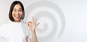 Everything okay. Smiling young asian woman assuring, showing ok sign with satisfied face, standing over white background