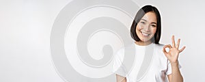 Everything okay. Smiling young asian woman assuring, showing ok sign with satisfied face, standing over white background