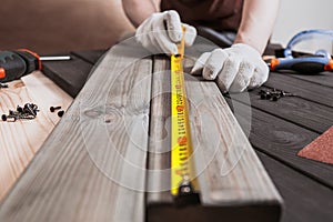 Exact measured. Close up of young male carpenter in gloves making measurements on the wooden plank by yellow measure tape