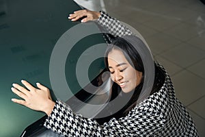 Exalted young asian woman hugging shining car hood