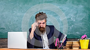 Examiner cunning face sit at table chalkboard background. Examinator bearded teacher with eyeglasses wondered face