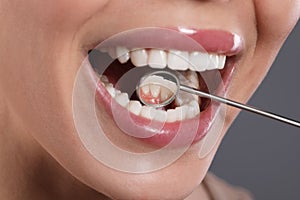 Examining woman`s teeth with dentist`s mirror on grey background, closeup