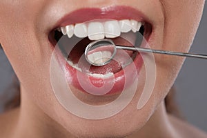 Examining woman`s teeth with dentist`s mirror on grey background, closeup