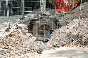 Excavation site for new drinking water pipe valve surrounded with protective metal fence or barrier on the sidewalk with pavement