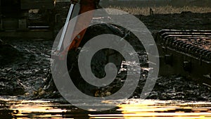 Excavator bucket digs soil from the bottom of the river at sunset. Cleaning and deepening the channel.