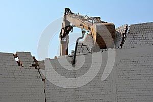 An excavator bucket is dismantling an old brick wall against the background of the sky