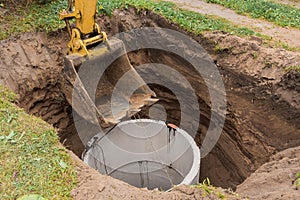 Excavator with a bucket, lowering into the pit on steel cables concrete sewer ring. Construction or repair of sewer home