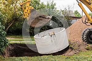 Excavator with a bucket, lowering into the pit on steel cables concrete sewer ring. Construction or repair of sewer home