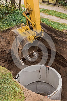 Excavator with a bucket, lowering into the pit on steel cables concrete sewer ring. Construction or repair of sewer home