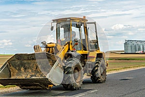 An excavator with a bucket rides along a rural road against the background of an agricultural field and a factory. A large modern