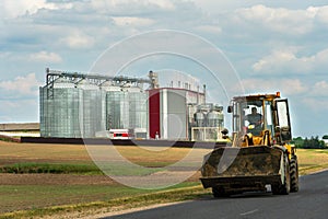 An excavator with a bucket rides along a rural road against the background of an agricultural field and a factory. A large modern