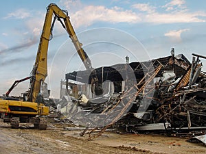 Excavator dismantling the charred remains of a burnt warehouse, twisted metal structures visible