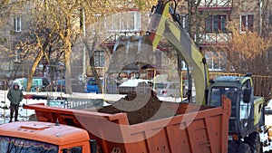 An excavator and a dump truck work on a construction site in winter. The excavator digs a hole