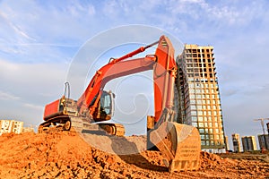 Excavator during excavation at construction site on sunset background. Red Backhoe on road work. Heavy Construction Equipment for