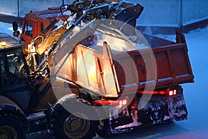 An excavator loads snow into the back of a dump truck at night.