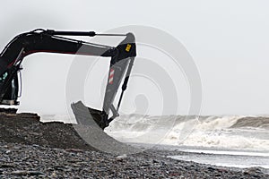 The excavator works with a bucket full of stones on seashore in stormy weather.