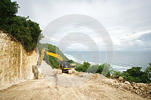 The excavator works on a rock cliff paving the way to the sea. Landscape with forest and ocean.