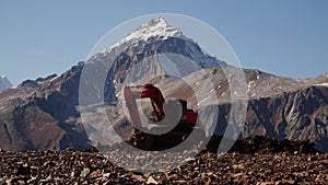 Excavator works in rocky Mountains. Creative. Excavator digs rocks at height of rocky mountains with snowy peaks