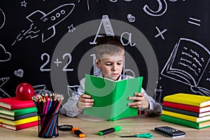 Excellent schoolboy sitting at the desk reading the book, surrounded with school supplies. Chalkboard as a background