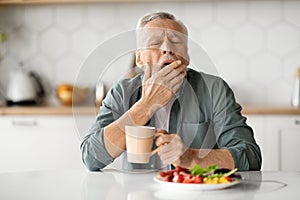 Excessive Daytime Sleepiness. Tired Senior Man Yawning At Table In Kitchen